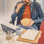 Male worker eating whole-grain popcorn while sitting at the computer with documents & mug| Corporate Synergies