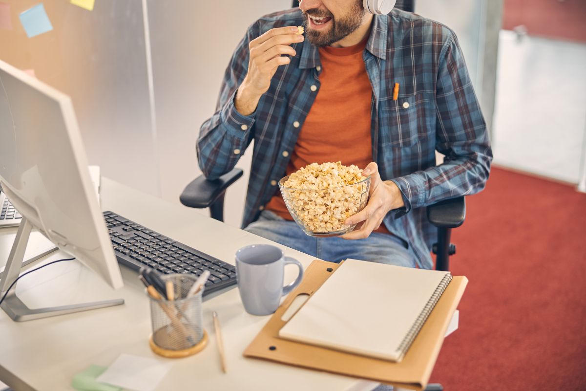 Male worker eating whole-grain popcorn while sitting at the computer with documents & mug| Corporate Synergies