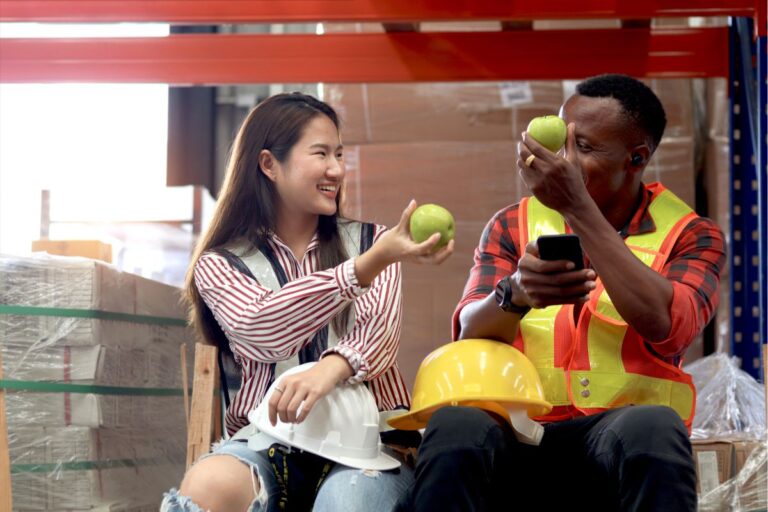 Asian female giving apple to African-American male co-worker in warehouse for heart health | Corporate Synergies