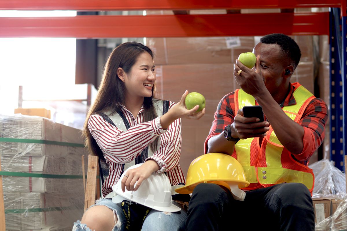 Asian female giving apple to African-American male co-worker in warehouse for heart health | Corporate Synergies