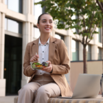 Happy businesswoman eating salad on bench outdoors for better nutrition | Corporate Synergies