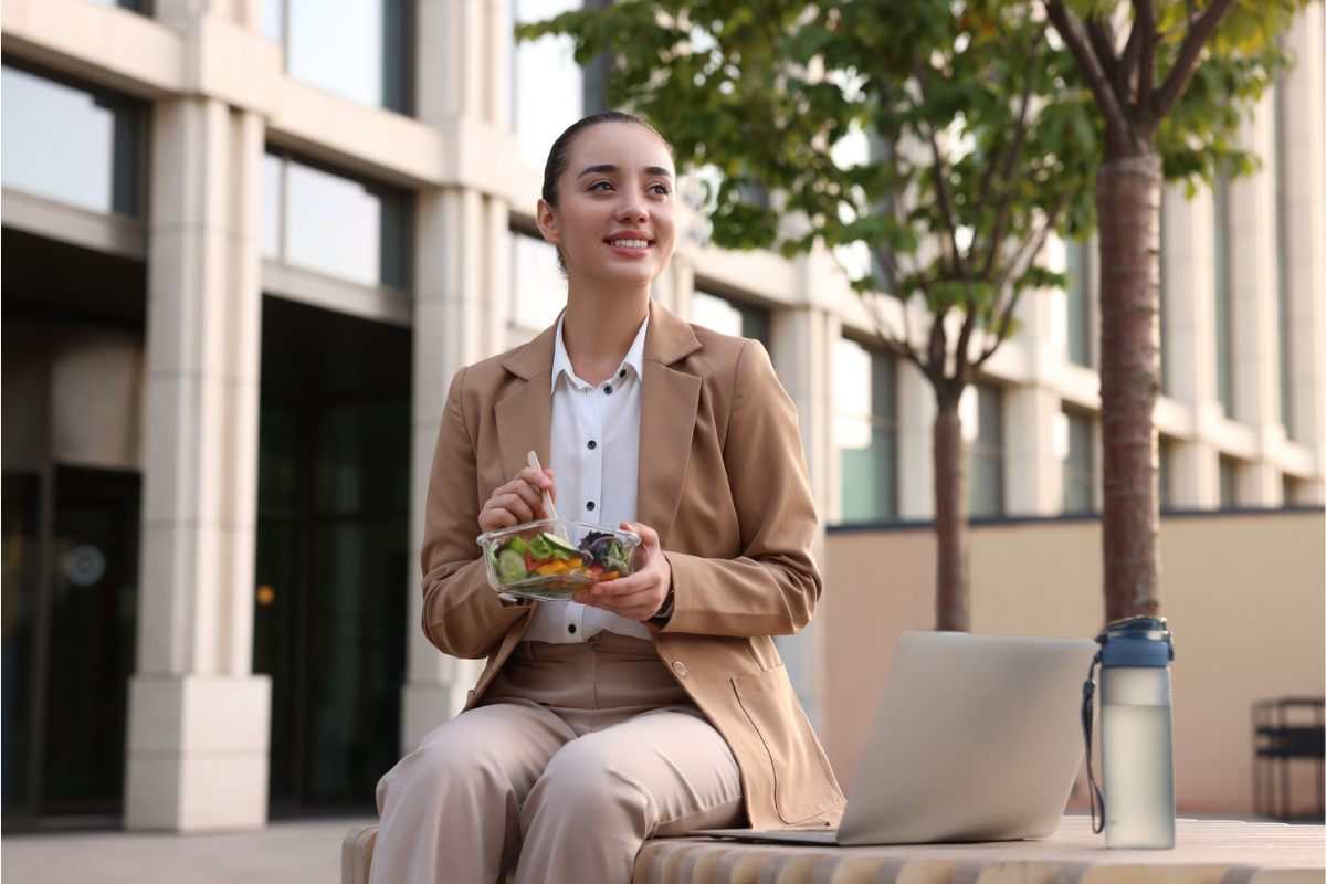 Happy businesswoman eating salad on bench outdoors for better nutrition | Corporate Synergies